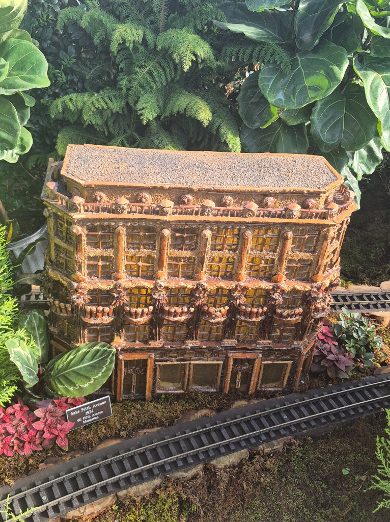 A miniature model of Saks Fifth Avenue made from natural materials, surrounded by lush green plants and flowers, with a small railway track nearby.